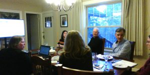 Seven people sitting around a table speaking and listening while a slideshow is playing on a large monitor in the background.