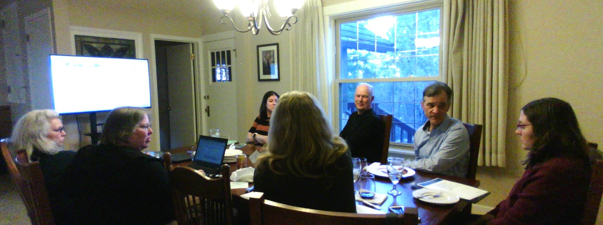 Seven people sitting around a table speaking and listening while a slideshow is playing on a large monitor in the background.